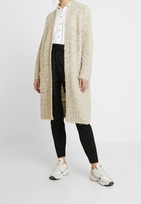 Lång beige cardigan med en texturerad, fluffig stickning. Bärs över en vit skjorta och svarta byxor, kombinerad med sportiga vita och rosa sneakers.