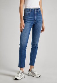 Blå denimjeans med hög midja, smal passform, rå fåll och femficksdesign, kombinerade med grå och vita mesh-sneakers.