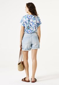 Camisa de manga corta con estampado floral, pantalones cortos de mezclilla azul claro, sandalias marrones y bolso de mimbre. Tejido suave, corte relajado, dobladillo enrollado en los pantalones cortos.