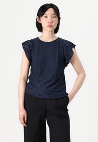Mørkeblå topp med korte volangermer, som har et teksturert øyelet-design med diamantmønstre. Brukt med svarte vide bukser.