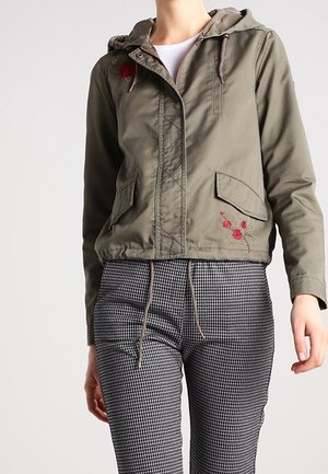 Osoba ubrana w oliwkowozieloną kurtkę z kapturem ozdobioną czerwonym kwiatowym haftem oraz w czarno-białe, w kratkę spodnie, stojąca na jednolitym tle.