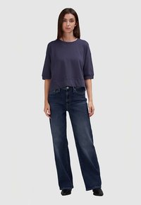 Marineblaues, kurzärmeliges cropped Sweatshirt kombiniert mit dunkelblauen, weit geschnittenen Jeans. Das Modell steht vor einem neutralen Hintergrund.