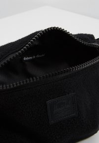 Czarny woreczek z polaru z zamkiem błyskawicznym, z gładką podszewką z tkaniny i czarną metką z logo wewnątrz. Zewnętrzna strona ma teksturowane wykończenie.