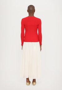 Haut rouge à manches longues en résille, avec un design ajusté, associé à une jupe maxi blanche fluide et des ballerines dorées, mettant en avant des textures lisses et des couleurs contrastées.