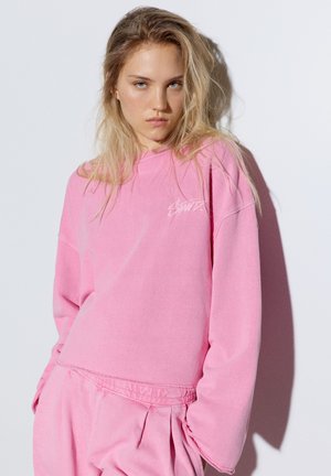 Ung kvinde med langt blondt hår iført stor pink sweatshirt og matchende bukser, der står mod en hvid væg med skygge.