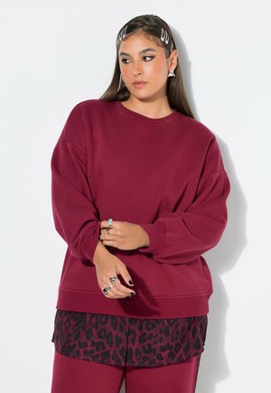 Kvinde med langt mørkt hår iført hårclips, en burgunder sweatshirt lag-på-lag over en sort og burgunder leopardprint skjorte og burgunder bukser.