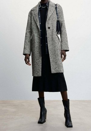 Długi płaszcz w szaro-czarną kratę w jodełkę, z klapowym kołnierzem i zapinany na pojedynczy guzik, zestawiony z czarną spódnicą i czarnymi botkami za kostkę.