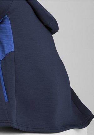 Vêtement bleu foncé avec un panneau latéral texturé bleu clair et une poche zippée, présenté sur un fond gris.