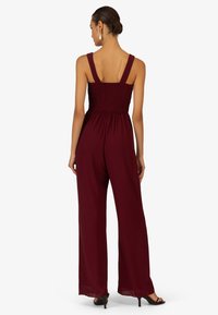 Burgunderfarbener Jumpsuit mit weitem Bein, gerafftem Oberteil und breiten Trägern. Aus leichtem Stoff gefertigt. Flache schwarze Absatzschuhe vervollständigen das Outfit.