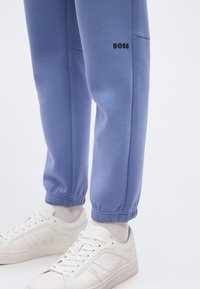 Πόδια με μπλε παντελόνια φόρμας με το λογότυπο «BOSS» πάνω από τα ελαστικά τελειώματα και λευκά αθλητικά παπούτσια με την επωνυμία «BOSS» στο πλάι.