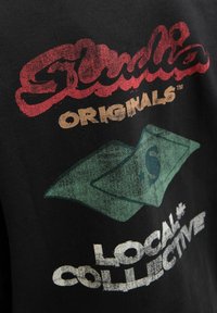 Čierne tričko s červeným nápisom "Studio Originals" a grafikou zelenej dolárovej bankovky. Obsahuje opotrebovaný biely nápis "Local Collective" nižšie.