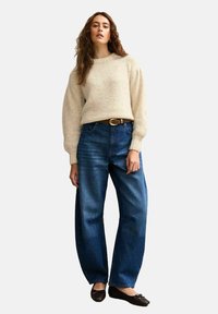 Crèmekleurige gebreide trui met lange mouwen, gecombineerd met hoog gewaagde blauwe spijkerbroek en zwarte platte schoenen.