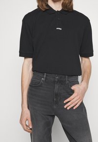 Polo shirt nera in cotone, con colletto classico, patta con tre bottoni e logo bianco del marchio. Abbinato a jeans grigio scuro.