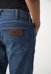 Jeanșii din denim bleu Wrangler prezintă o etichetă din piele pe spate cu detalii cusute și un croi slim, evidențiind un design clasic cu cinci buzunare.