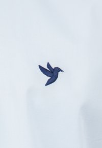 Colibrí bordado en azul sobre una tela azul claro, que presenta un diseño sencillo con detalles en el bordado y una textura suave.