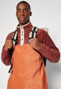Fleece-pullover i mörkröd med hög krage och knappstängning, lager över en orange fleeceförkläde. Ryggsäcksremmar i ljusgrå.