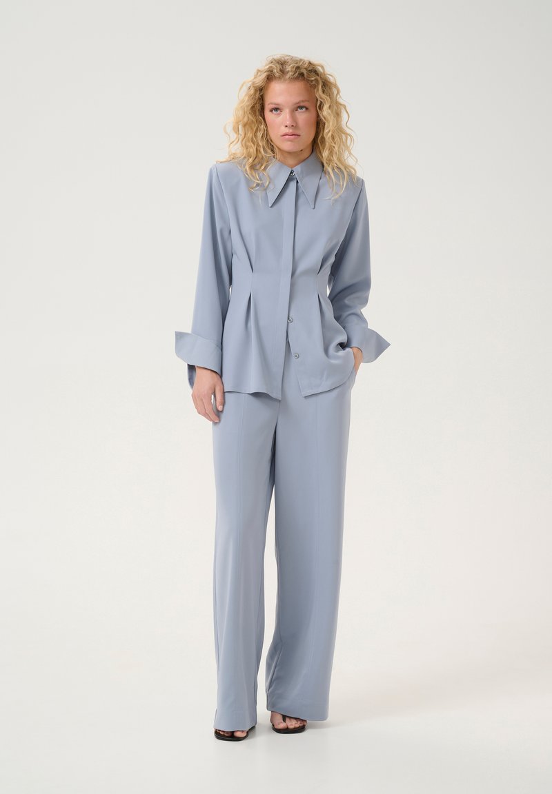 Completo su misura azzurro chiaro con camicia a bottone caratterizzata da un ampio colletto e pieghe, abbinato a pantaloni a gamba larga e sandali con tacco.