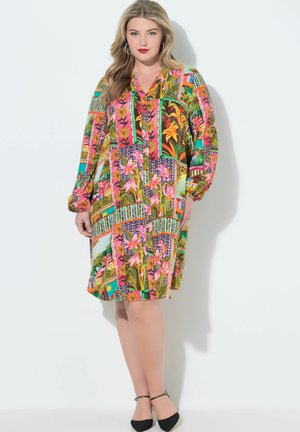 Kobieta ubrana w kolorową sukienkę patchworkową do kolan z długimi rękawami i czarne czółenka na pasku wokół kostki, stojąca na białym tle.