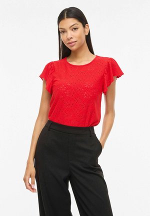 Kvinde iført en rød top med korte ærmer og hulmønster samt sorte, højtaljede bukser, der står med den ene hånd i lommen mod en hvid baggrund.