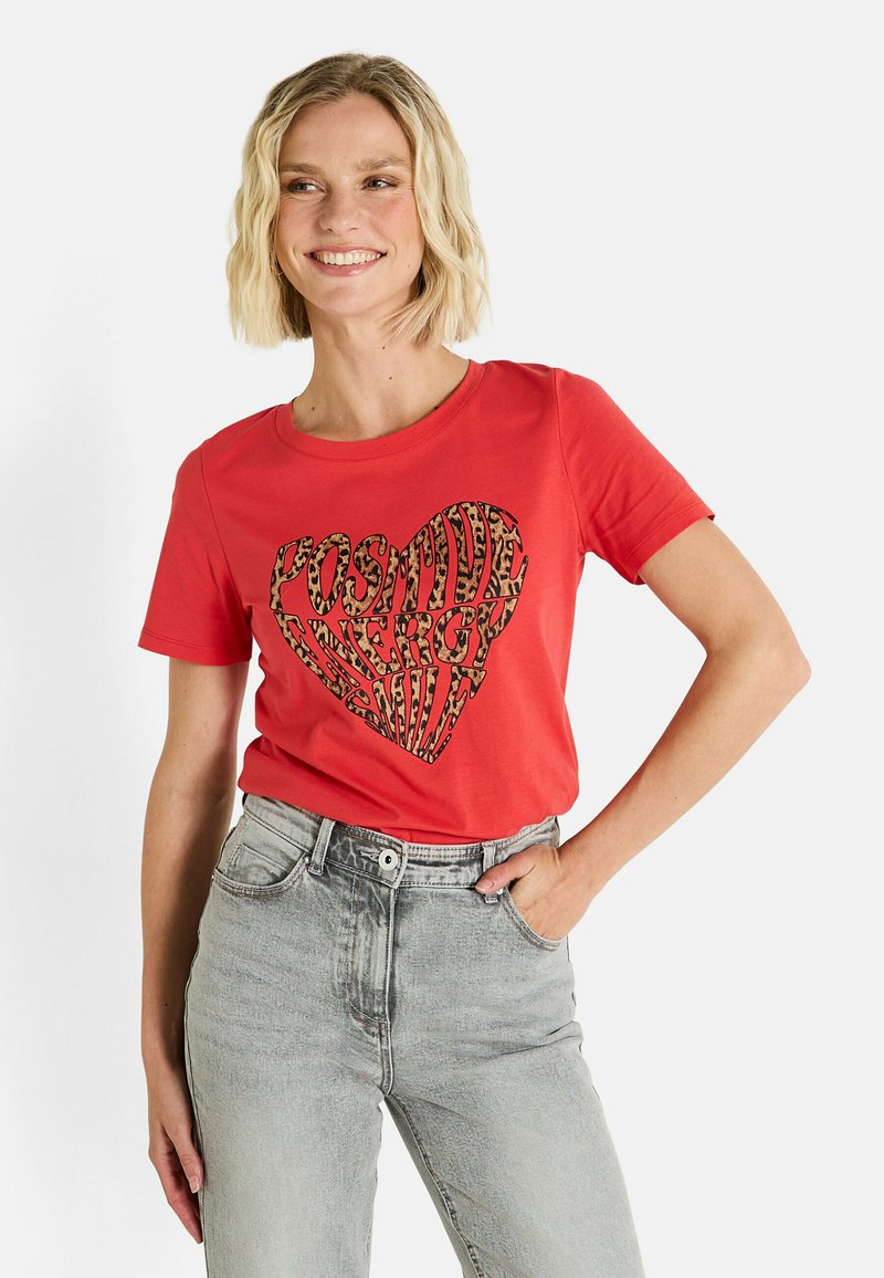 Camiseta de algodón roja con cuello redondo, que presenta un diseño de corazón con estampado de leopardo y texto en negro que dice "ENERGÍA POSITIVA SONRÍE."