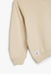 Sudadera beige con puños y dobladillo acanalados, con un acabado texturizado y una pequeña etiqueta de logo en el borde inferior.