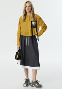 Camisa abotonada mostaza con un bolsillo gráfico, combinada con una falda gris oscuro con ribete blanco. Bolsa negra con acento amarillo y zapatillas grises.
