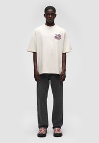 Overdimensioneret cremet t-shirt med et burgunder grafisk logo på brystet, parret med mørkegrå bukser og mønstrede sko. Enkel design, afslappet pasform.