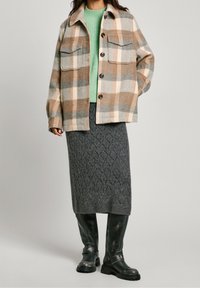 Mujer vistiendo una chaqueta de cuadros en beige y gris sobre un suéter verde menta, falda de punto gris y botas negras hasta la rodilla.