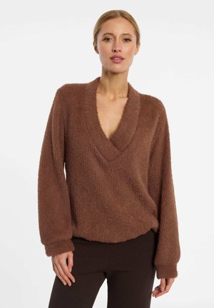 Femme portant un pull marron à col en V duveteux à manches longues et un pantalon foncé, debout devant un fond clair uni.
