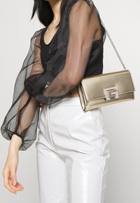 Sac à main en métal doré avec une bandoulière chaîne, orné d'un fermoir logo bijoux. Le mannequin porte des manches noires transparentes et un pantalon blanc ajusté.