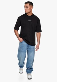 Black cotton t-shirt with a round neck, short sleeves, and a rectangular gray label at the chest, paired with light blue baggy jeans and white sneakers.