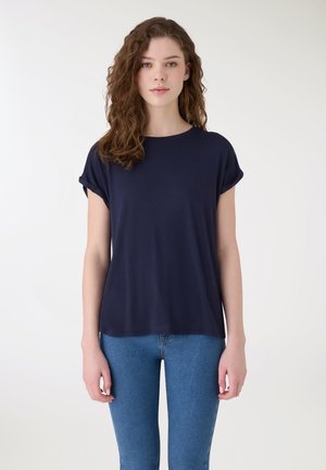 Giovane donna con capelli castani ondulati, che indossa una maglietta blu navy a maniche corte e jeans aderenti, in piedi davanti a uno sfondo bianco.