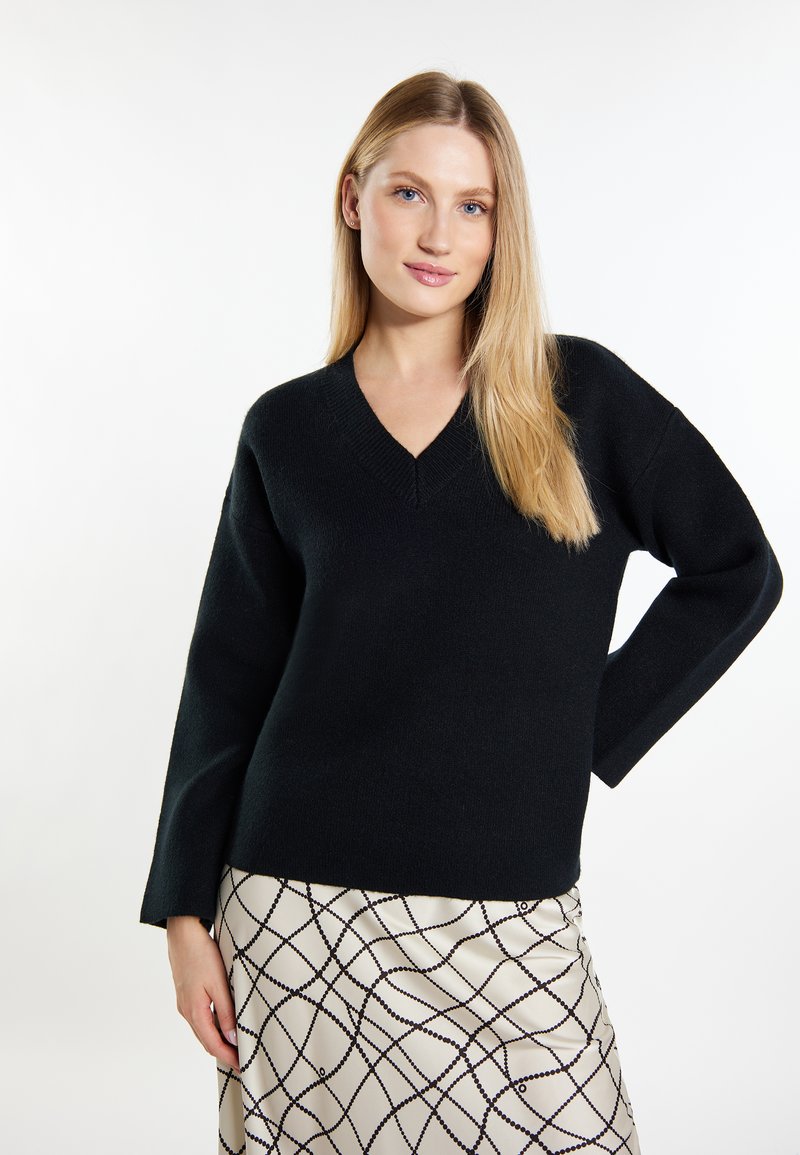 Schwarzer V-Ausschnitt-Pullover mit langen Ärmeln, aus einem weichen Stoff; kombiniert mit einem glatten cremfarbenen Rock mit einem Kreuzmuster.