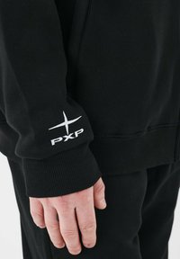 Sort sweatshirt med ribbede håndled og kant. Har en hvid "PXP" logo brodering på ærmet. Blød stof tekstur.