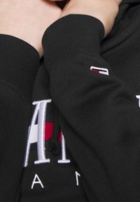 Sudadera negra con puños de punto, que presenta un gran logotipo bordado en blanco con acentos en rojo y azul en la parte delantera y un pequeño logotipo en la manga.