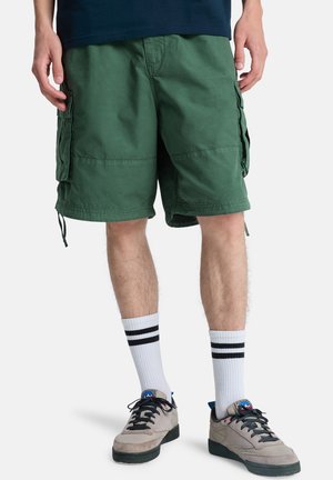Groene cargoshorts met zijzakken en trekkoorden, gemaakt van lichtgewicht stof. Gecombineerd met witte sokken en grijze sneakers.