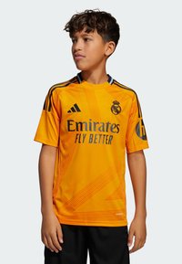 Maillot de football orange à manches courtes, avec des accents noirs et le logo du Réal Madrid, portant le texte "Emirates Fly Better".