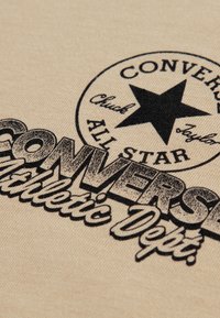 Μπεζ ύφασμα με μαύρο τυπωμένο λογότυπο που περιλαμβάνει ένα αστέρι και το κείμενο "CONVERSE ALL STAR Athletic Dept." σε ρετρό γραμματοσειρά.