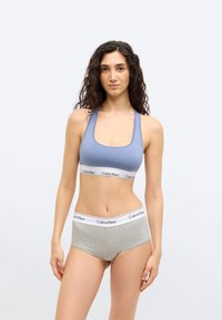 Νεαρή γυναίκα με σγουρά σκούρα μαλλιά, που φοράει μπλε αθλητικό σουτιέν και γκρι εσώρουχα Calvin Klein, στέκεται μπροστά σε λευκό φόντο.