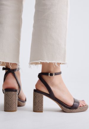 Pies con vaqueros beige recortados y deshilachados y sandalias marrones de tacón de bloque con tiras en el tobillo y tacones con patrón tejido sobre fondo blanco.