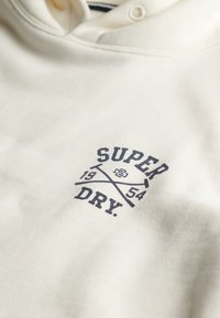 Kremowa bluza z kapturem wykonana z miękkiego materiału, z ciemnoniebieskim nadrukowanym logo z krzyżującymi się liniami oraz napisem "SUPER DRY" i rokiem "1954."