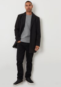 Sort uldfrakke med slagkrave over en lys grå ribbet sweater, parret med smalle sorte bukser og sorte sneakers. Ensfarvet baggrund.