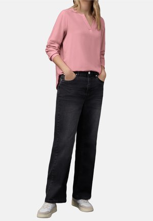 Kvinde iført en pink bluse med lange ærmer, sorte vidde jeans og hvide sneakers, stående med hænderne i lommerne foran en ensfarvet baggrund.