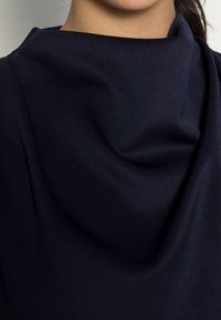 Kvinna som bär en mörk marinblå topp med en hög, lös krage som visar halsen och delar av käklinjen.