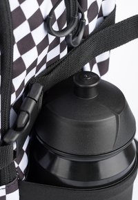 Bottiglia d'acqua nera con tappo a vite, poggiata su una tracolla nera testurizzata contro una borsa a righe bianche e nere.
