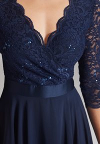 Marineblaues Kleid mit Spitzenoberteil, gewelltem Ausschnitt und Paillettenakzenten. Figurbetonter Taillenbereich mit einem glatten Rock, der unterhalb der Hüften fließt.