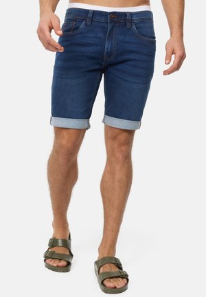 Shorts en denim bleu foncé, avec des ourlets roulés et un design classique à cinq poches. Associés à des sandales vert olive.