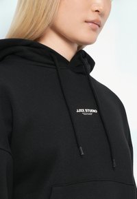 Svart hettegenser laget av mykt materiale med kengurulomme, med en hvit logo på brystet og svarte snorer til hetten.