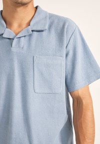 Camisa polo de color azul claro hecha de tejido texturizado, con cuello en punta y un bolsillo en el pecho izquierdo. Diseño de mangas cortas y ajuste holgado.