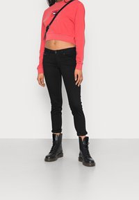 Felpa rossa cropped con logo, abbinata a jeans skinny neri e stivali stringati neri. L'insieme presenta un design casual e aderente.